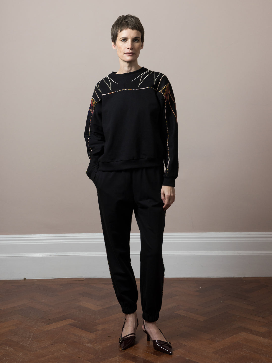 model standing on parquet floor wears black sweatshirt in cotton fleece with artisan embroidery across arms and shoulders with matching  Joggers black cotton fleece with piping detail