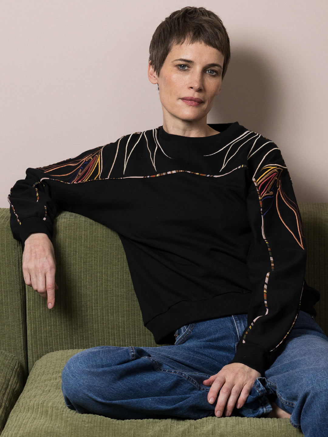 model sitting on green sofa wears black sweatshirt in cotton fleece with artisan embroidery across arms and shoulders with jeans