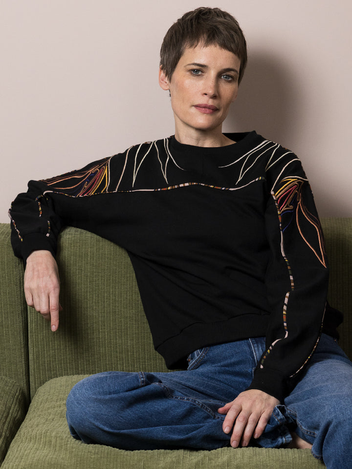 model sitting on green sofa wears black sweatshirt in cotton fleece with artisan embroidery across arms and shoulders with jeans