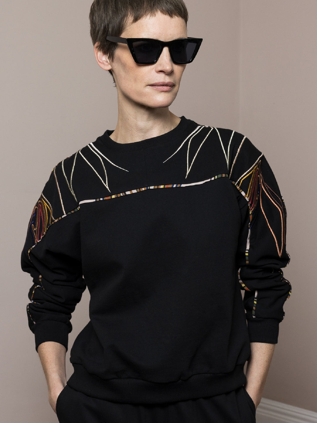 model wears black sweatshirt in cotton fleece with artisan embroidery across arms and shoulders with matching joggers hands in pocket wearing sunglasses