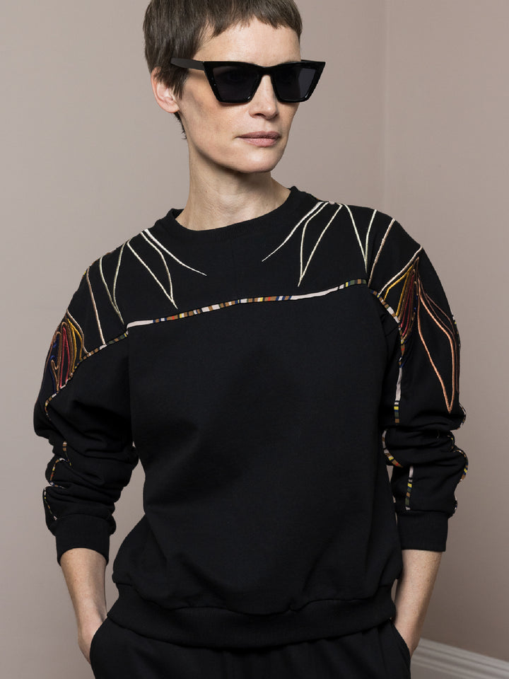 model wears black sweatshirt in cotton fleece with artisan embroidery across arms and shoulders with matching joggers hands in pocket wearing sunglasses