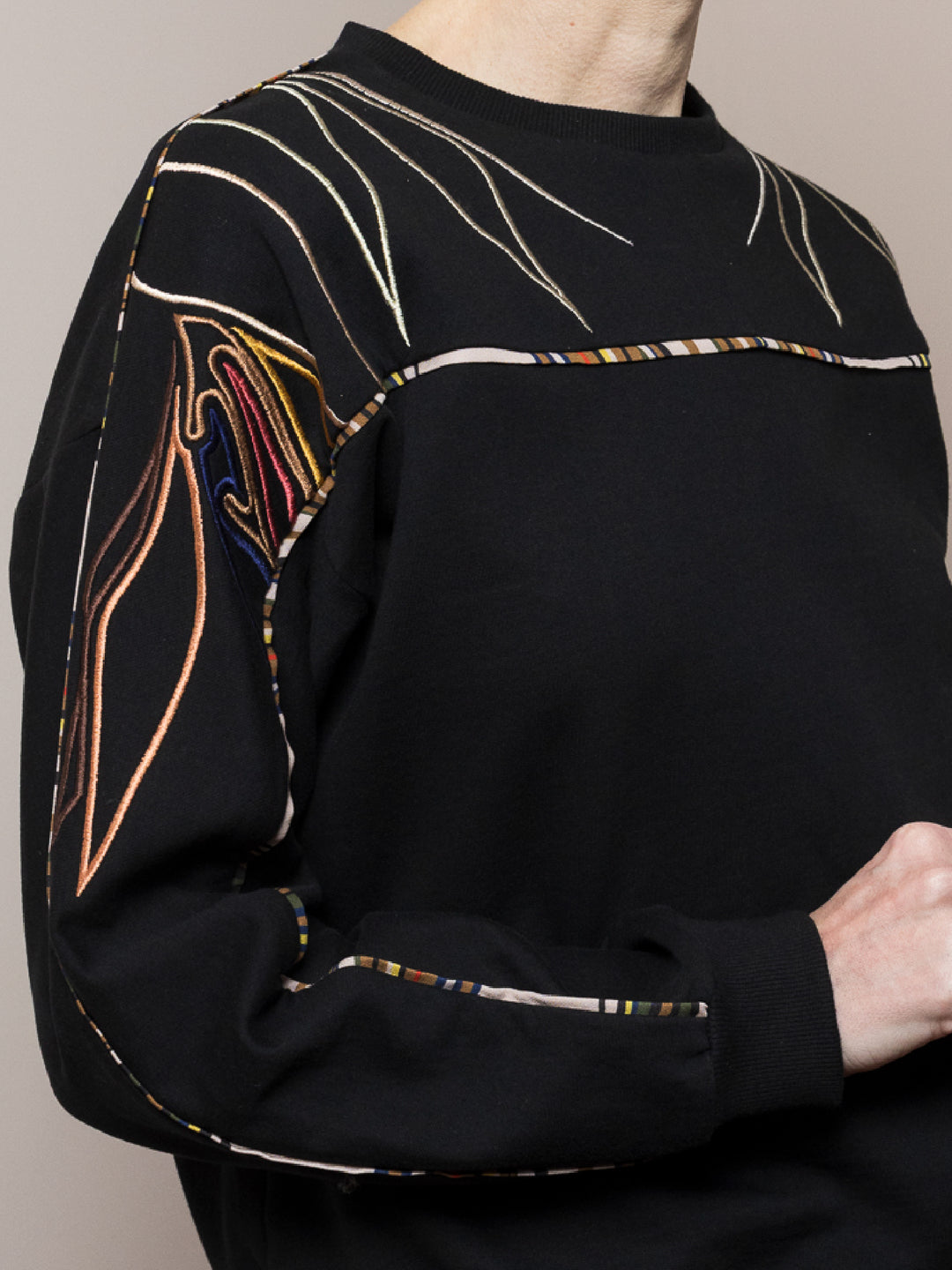 close of model standing  wears black sweatshirt in cotton fleece with artisan embroidery across arms and shoulders il