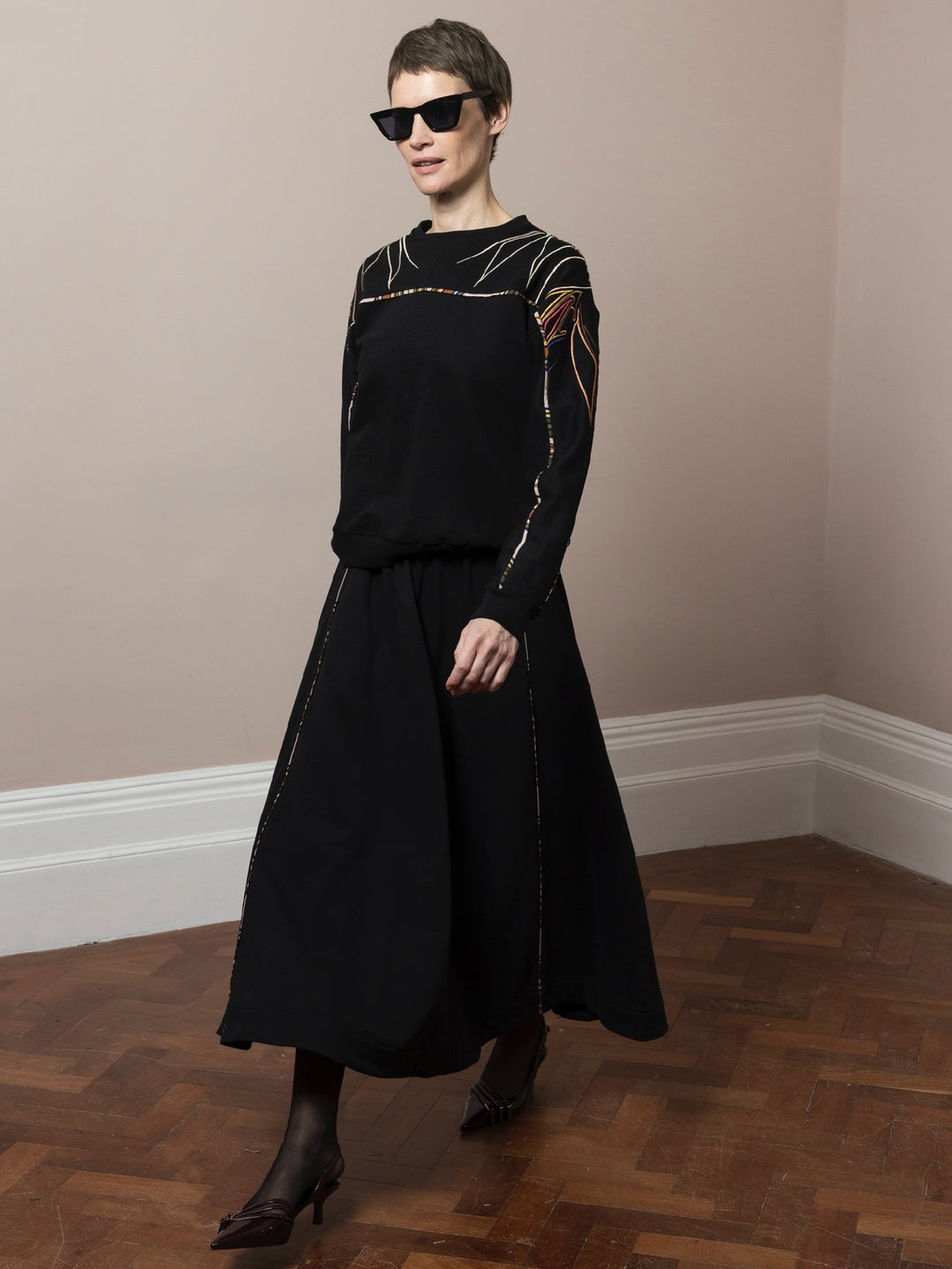 model wearing black skirt in cotton fleece with piping walking and co-ord Sweatshirt in black cotton fleece with embroidery across arms and shoulders walking on parquet floor wearing sunglasses