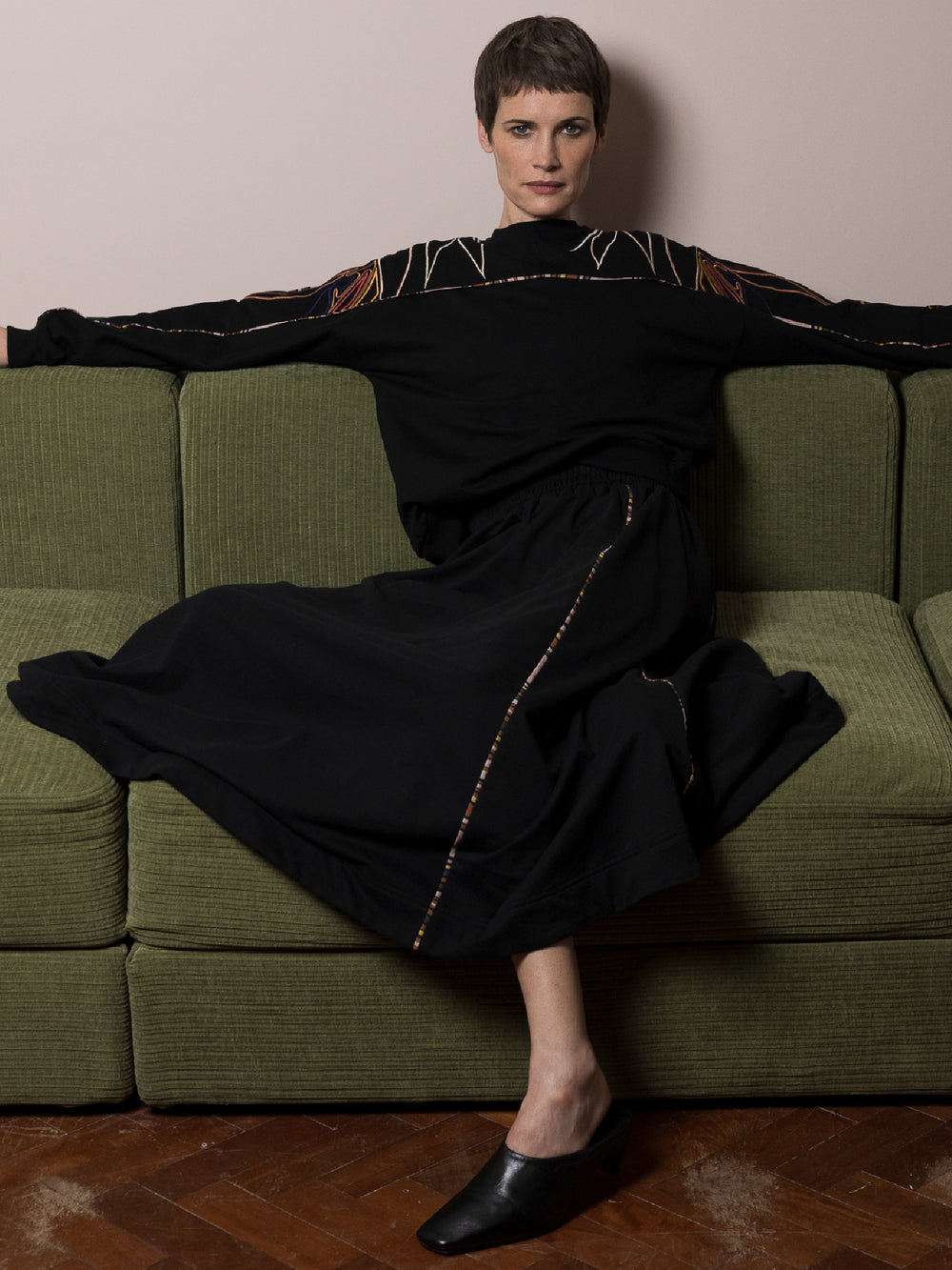 model wearing black skirt in cotton fleece with piping walking and co-ord Sweatshirt in black cotton fleece with embroidery across arms and shoulders sitting on green sofa in living room 