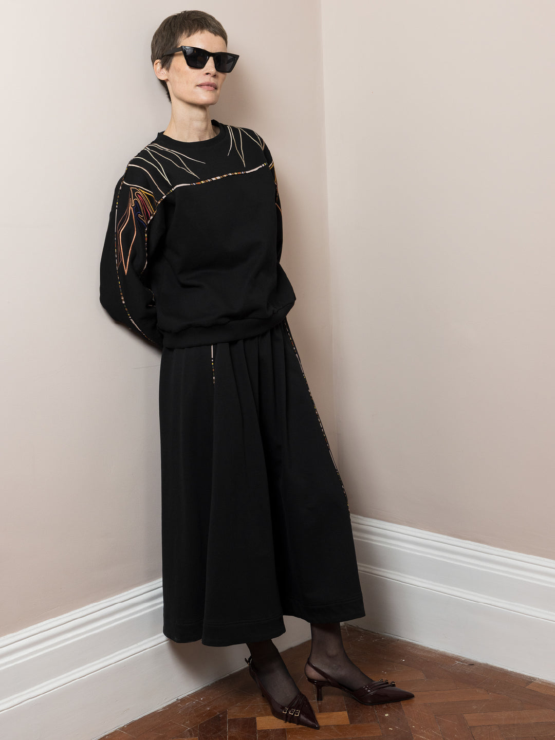 model leaning on wall wearing black skirt in cotton fleece with piping walking and co-ord Sweatshirt in black cotton fleece with embroidery across arms and shoulders model standing on parquet floor wearing sunglasses