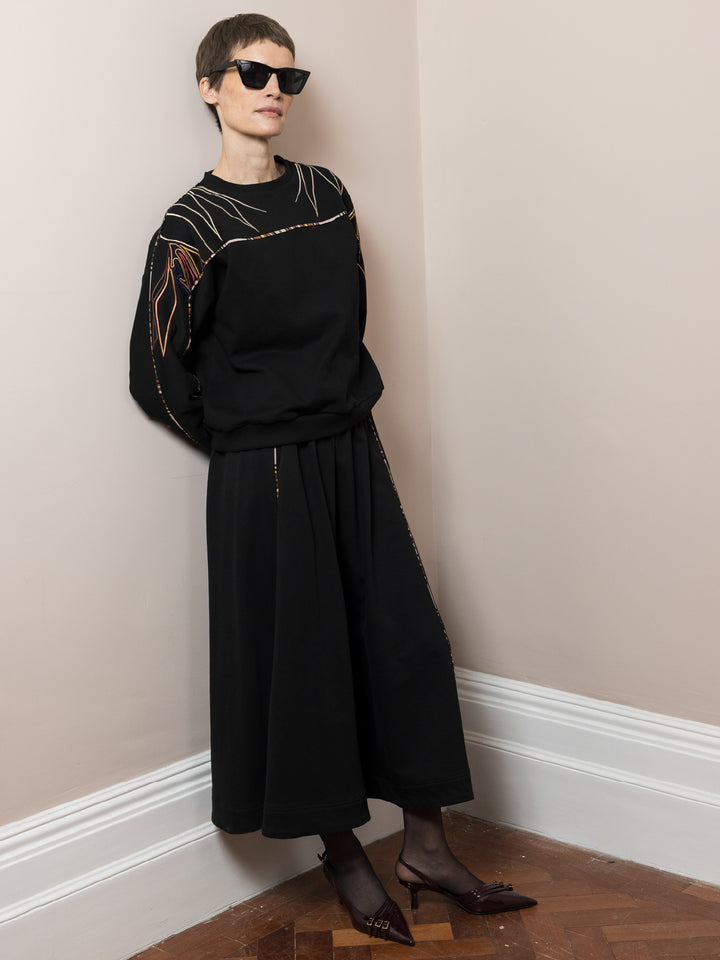model leaning on wall wearing black skirt in cotton fleece with piping walking and co-ord Sweatshirt in black cotton fleece with embroidery across arms and shoulders model standing on parquet floor wearing sunglasses