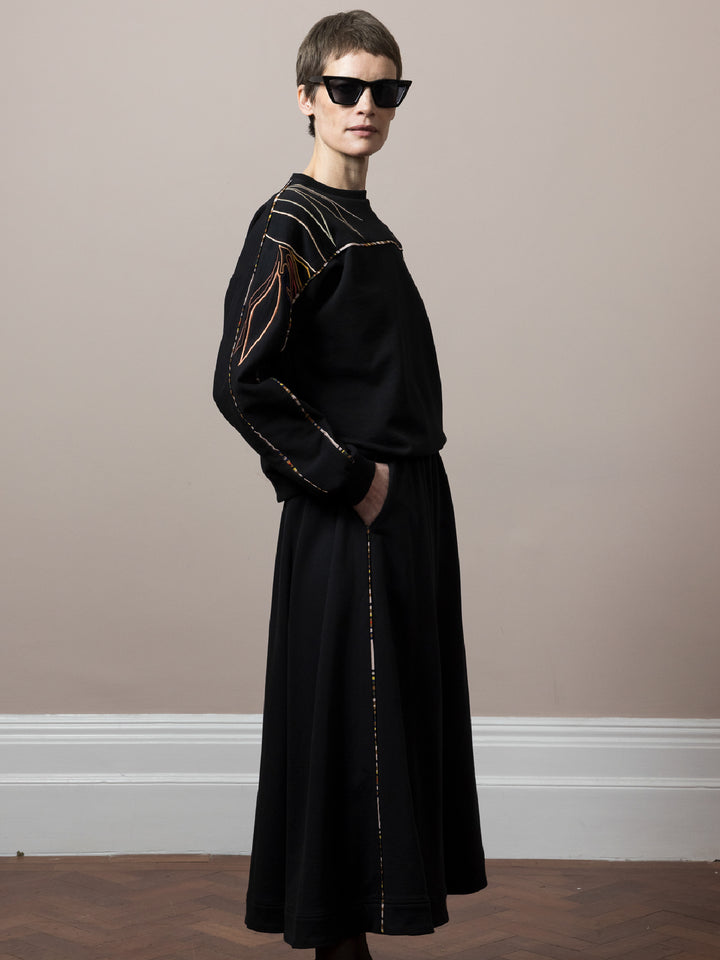 model standing on parquet floor wears black sweatshirt in cotton fleece with artisan embroidery across arms and shoulders with matching skirt with piping detail 