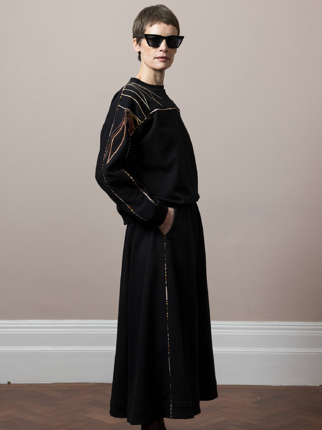 model wearing black skirt in cotton fleece with piping walking and co-ord Sweatshirt in black cotton fleece with embroidery across arms and shoulders standing on parquet floor showing side view with hands in skirt pocket wearing sunglasses