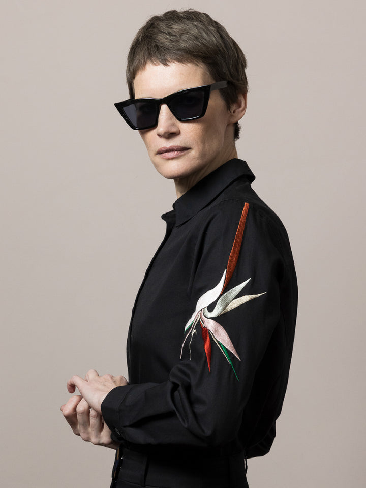 model wears Black shirt in cotton satin poplin with artisan embroidery on the arm looking at camera wearing black sunglasses