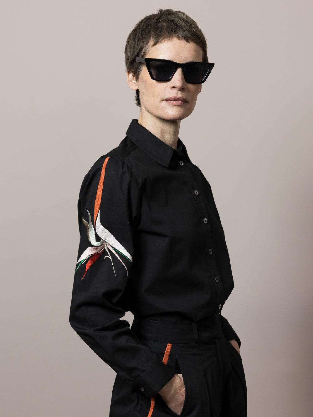 model wears Black shirt in cotton satin poplin with artisan embroidery on the arm styled with matching black cotton trousers  wearing black sunglasses