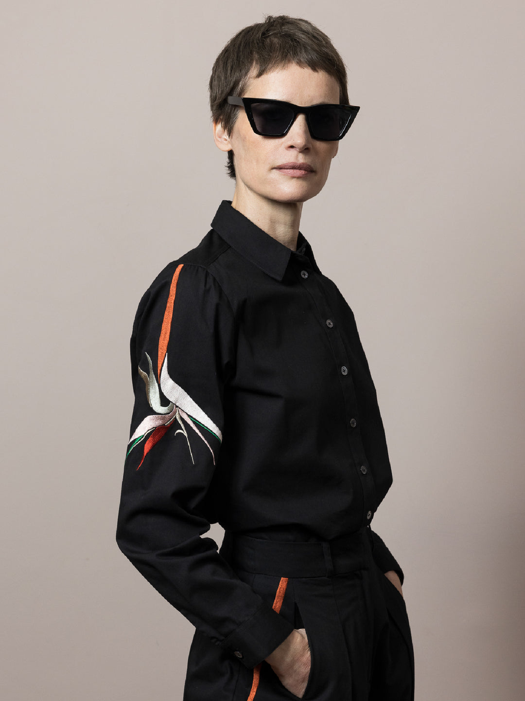 model wears Black shirt in cotton satin poplin with artisan embroidery on the arm styled with matching black cotton trousers  wearing black sunglasses
