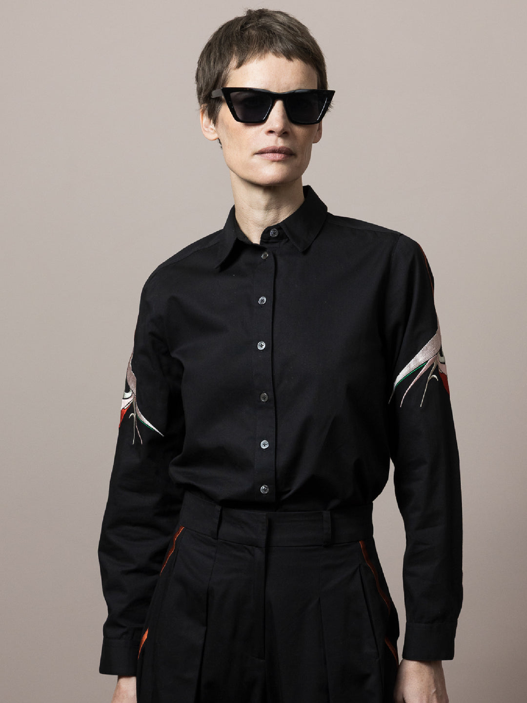 front view of model wears Black shirt in cotton satin poplin with artisan embroidery on the arm styled with matching black cotton trousers  wearing black sunglasses