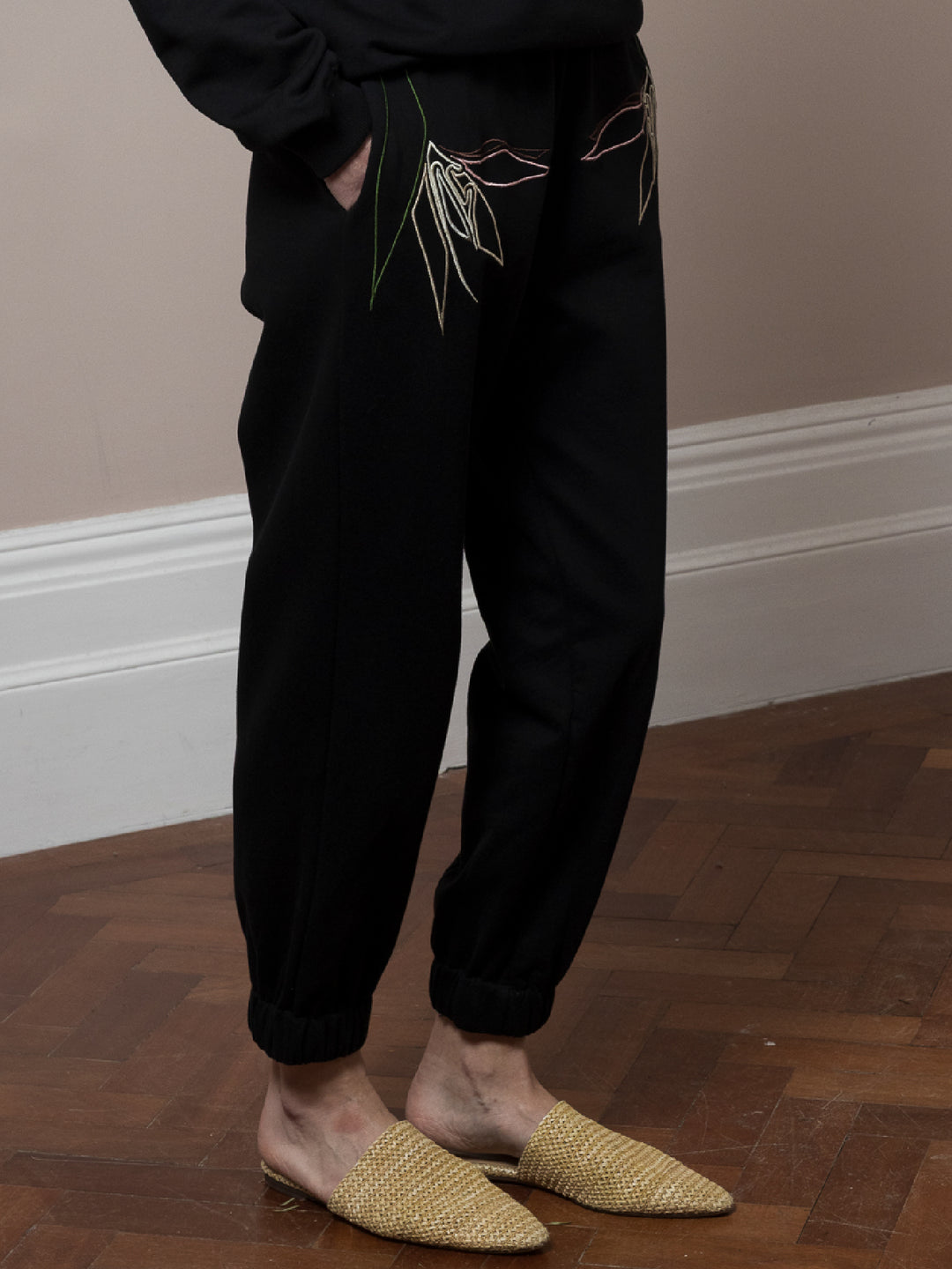 model standing on parquet floor with hands in pockets wears Black Joggers with artisan embroidery across pockets in cotton fleece 