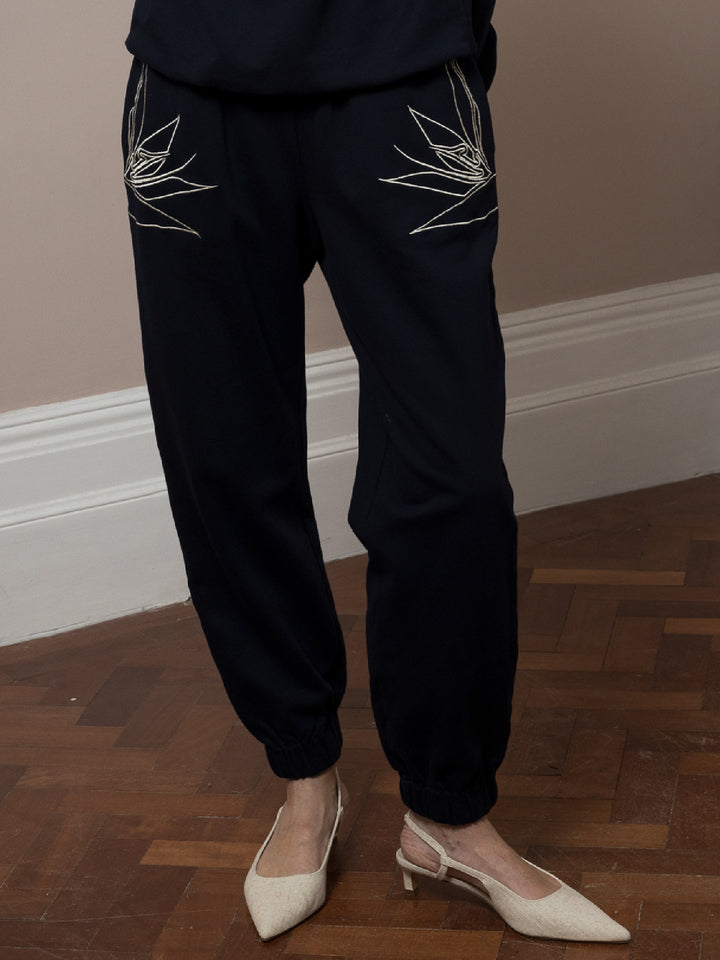 view of model  standing on parquet floor wears  dark navy Joggers with artisan cream linear embroidery across pockets in cotton fleece 