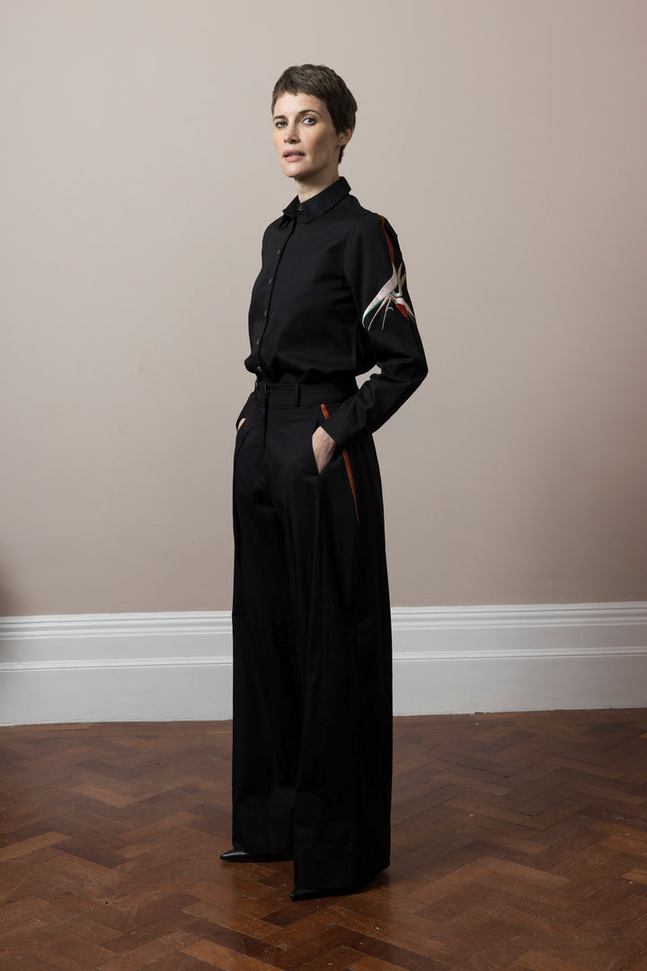 Model wears wide leg trousers black cotton satin poplin with embroidery detail with matching Shirt black cotton poplin embroidered standing on parquet wooden floor