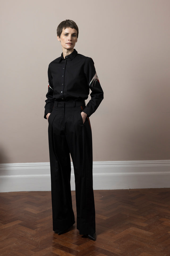 model wearing co-ord outfit of black cotton satin trousers and blouse both embroidered in Mumbai by artisans standing on wooden parquet floor  against pale pink wall 