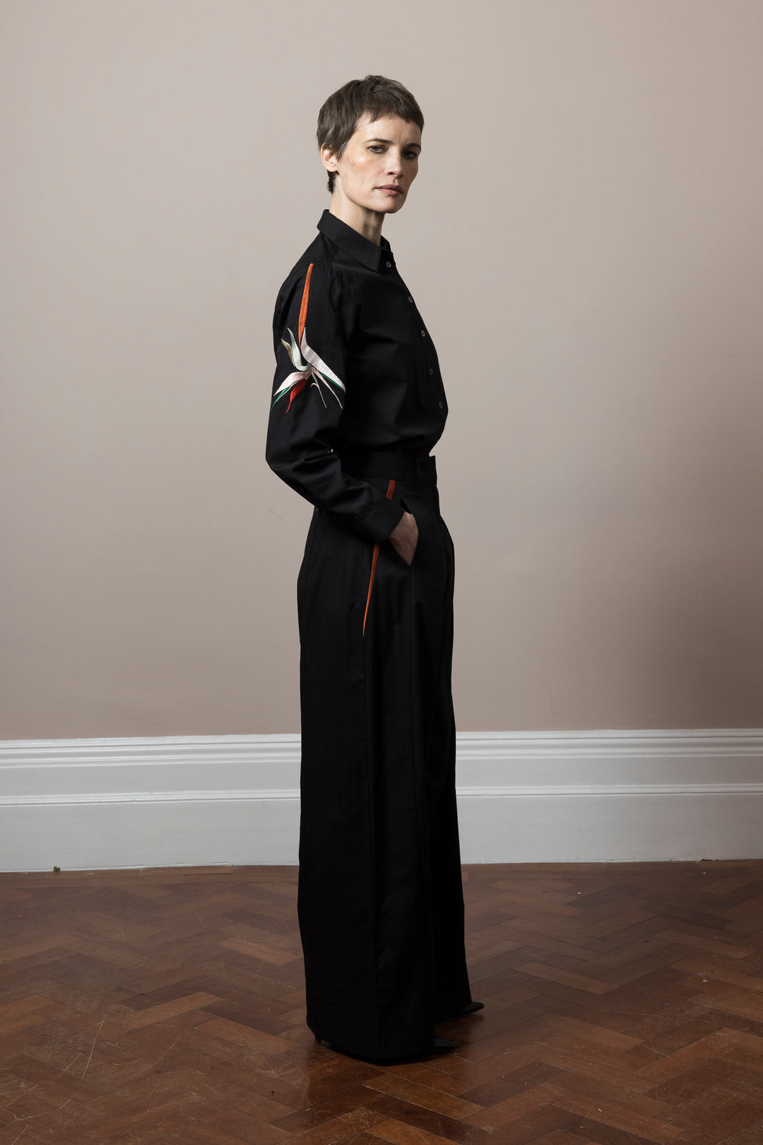 model wearing co-ord outfit of black cotton satin trousers and blouse  side view embroidery done in Mumbai by artisans model is tanding on wooden parquet floor  against pale pink wall showing side view 