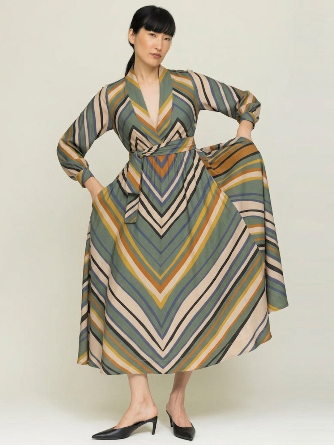 model wearing stripe print wrap dress with  hands in pockets showing wide skirt against a neutral background 
