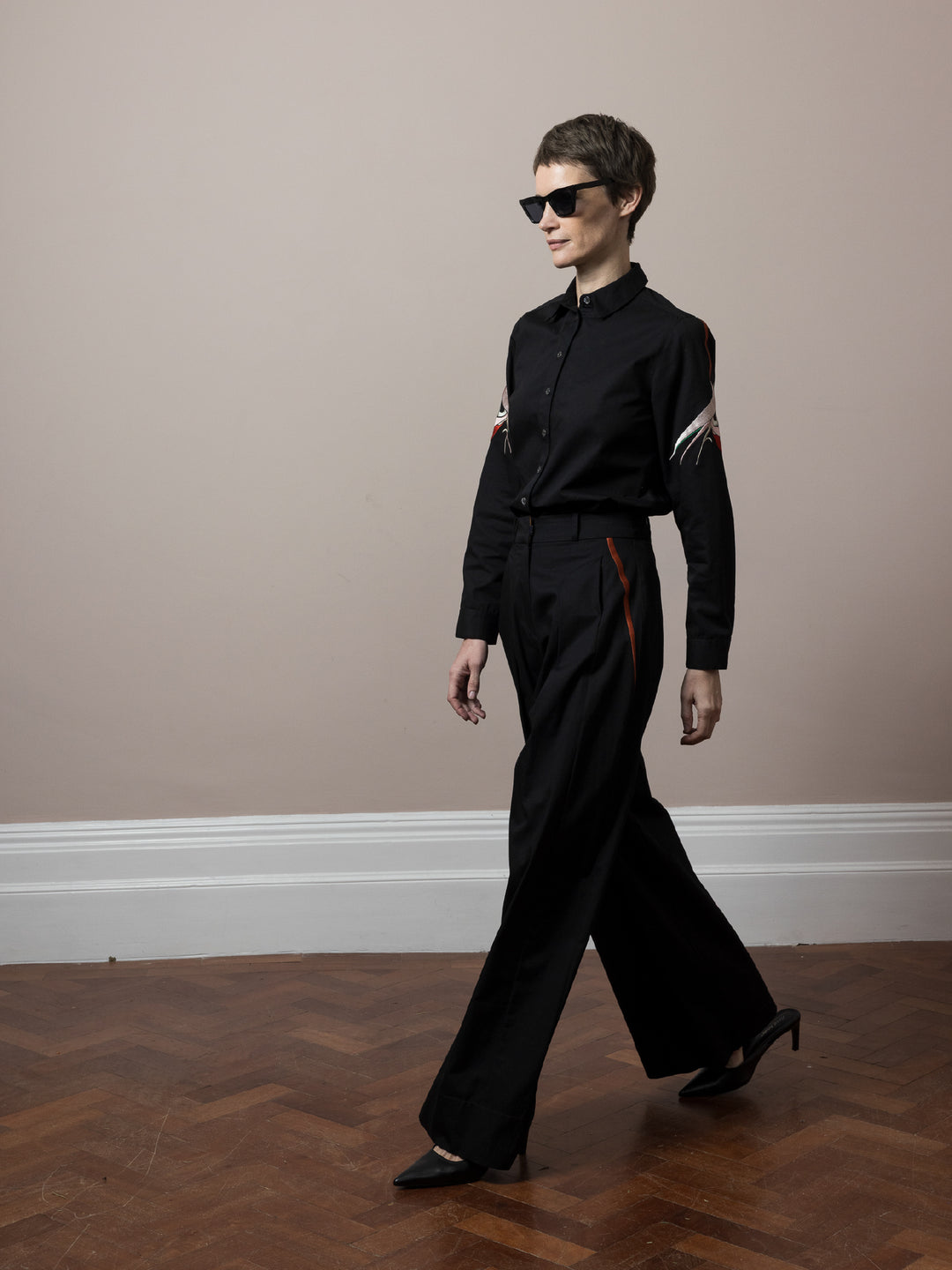 model wearing co-ord outfit of black cotton satin trousers and blouse both embroidered in Mumbai by artisans walking on wooden parquet floor wearing sunglasses 