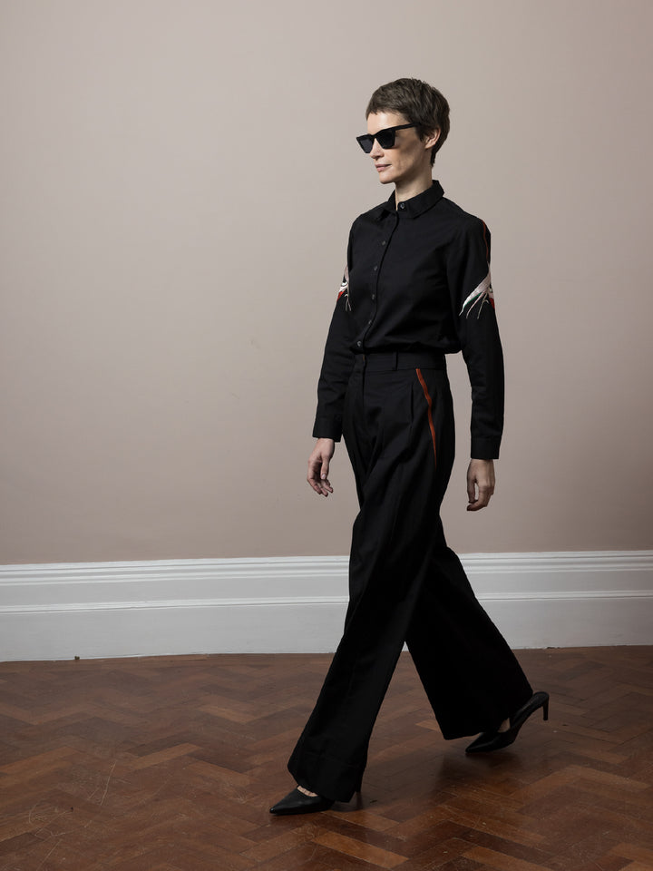 model wearing co-ord outfit of black cotton satin trousers and blouse both embroidered in Mumbai by artisans walking on wooden parquet floor wearing sunglasses 