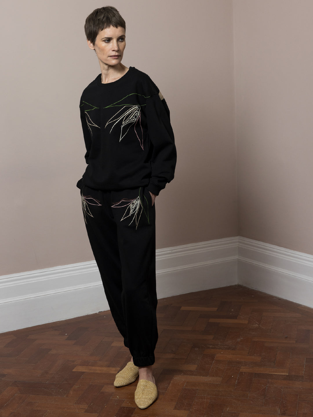 view of model  standing on parquet floor wears  Black sweatshirt cotton fleece with linear embroidery across front and artisan chainstitch embroidery on back with matching black Joggers with artisan linear embroidery across pockets in cotton fleece with her hands in pockets