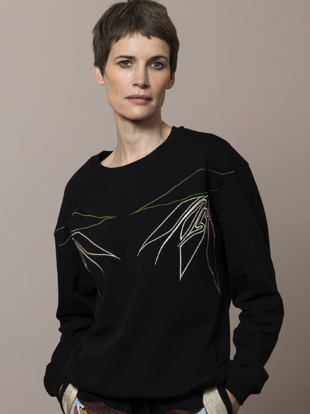 view of model  wears  Black sweatshirt cotton fleece with linear embroidery across front and artisan chainstitch embroidery on back with matching black Joggers hands in pockets with artisan chainstitch embroidery across pockets in cotton fleece 