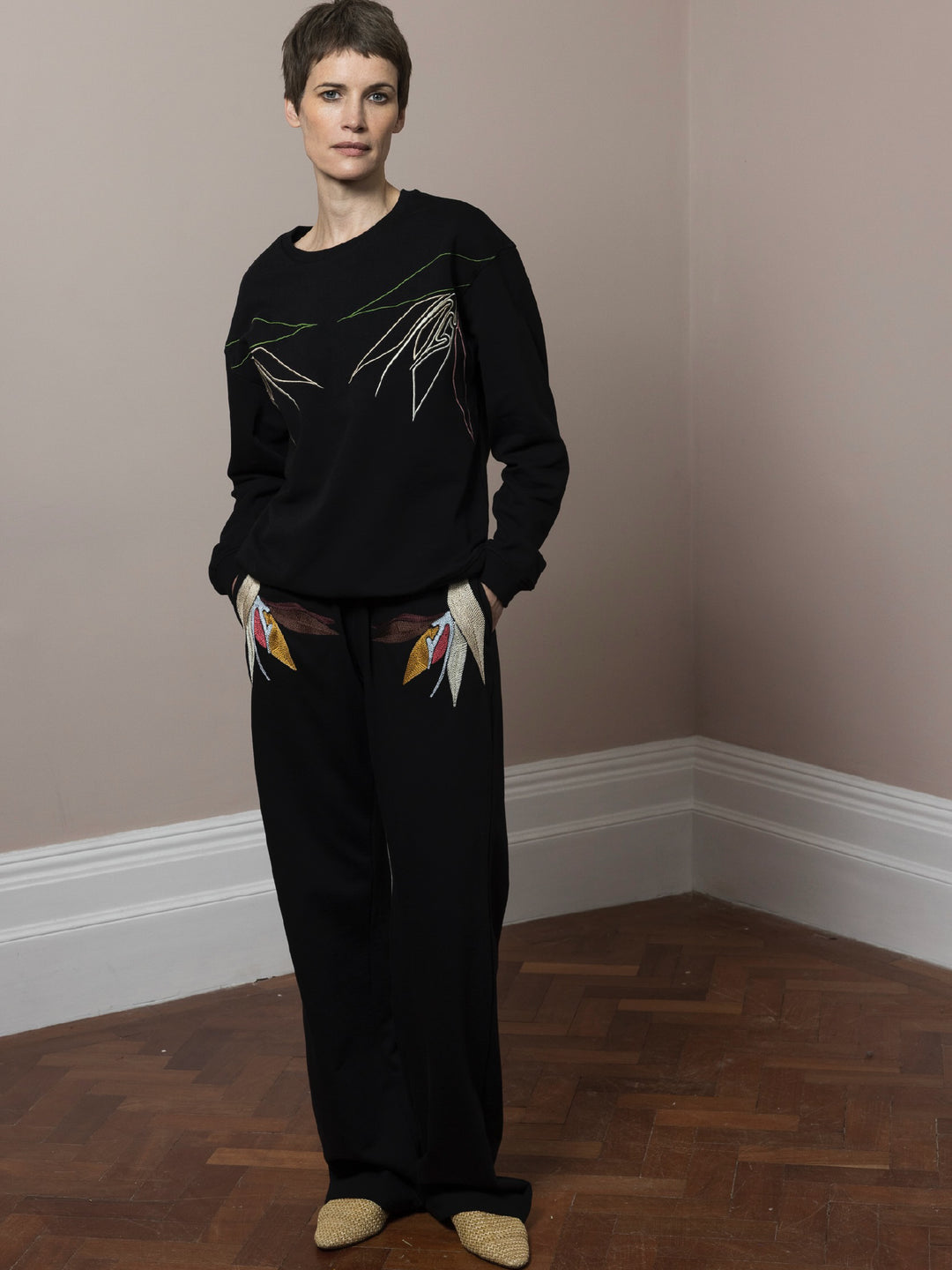 model standing on parquet floor with hands in pockets wears  Black sweatshirt cotton fleece with linear embroidery across front and chainstitch embroidery on back with matching Joggers with artisan embroidery across pockets in cotton fleece 