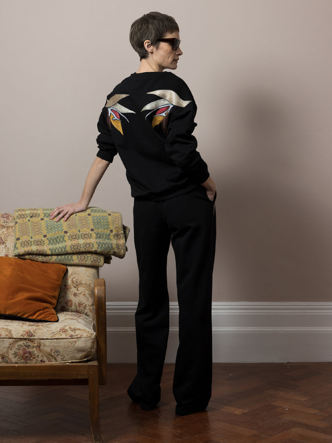view of model leaning on sofa on parquet floor wears  Black sweatshirt cotton fleece with linear embroidery across front and artisan chainstitch embroidery on back with matching black Joggers in cotton fleece 