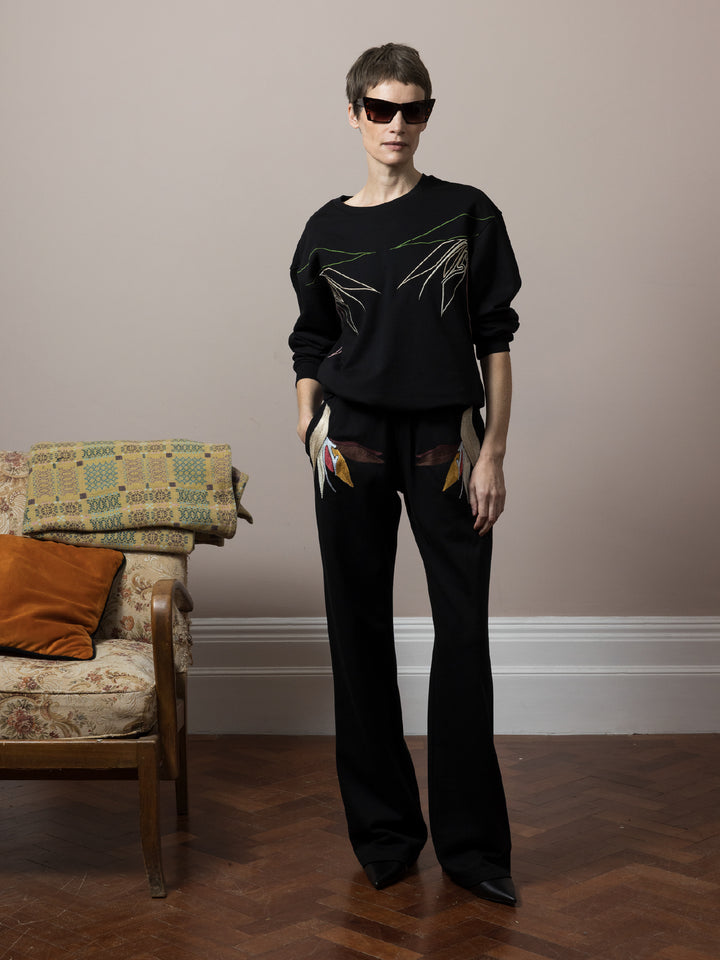 model standing by sofa on parquet floor with hands in pockets wears  Black sweatshirt cotton fleece with linear embroidery across front and chainstitch embroidery on back with matching Joggers with artisan embroidery across pockets in cotton fleece 
