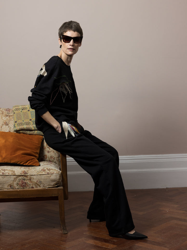 model leaning on sofa on parquet floor with hands in pockets wears  Black sweatshirt cotton fleece with linear embroidery across front and chainstitch embroidery on back with matching Joggers with artisan embroidery across pockets in cotton fleece 