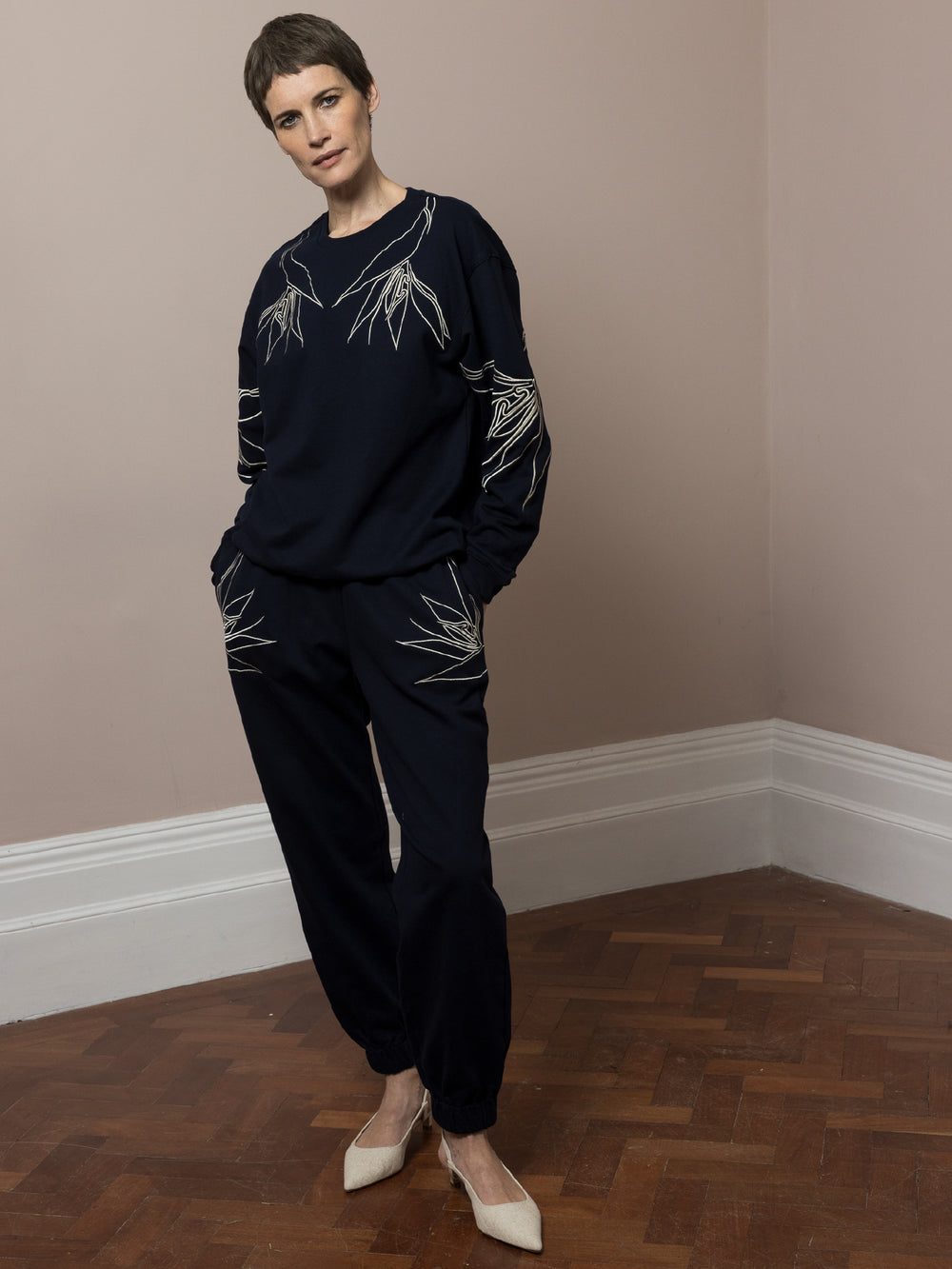 view of model  standing  with hands in pockets on parquet floor wears  dark navy Joggers with artisan cream linear embroidery across pockets in cotton fleece with matching sweatshirt in dark navy with cream artisan linear embroidery across front and arms 