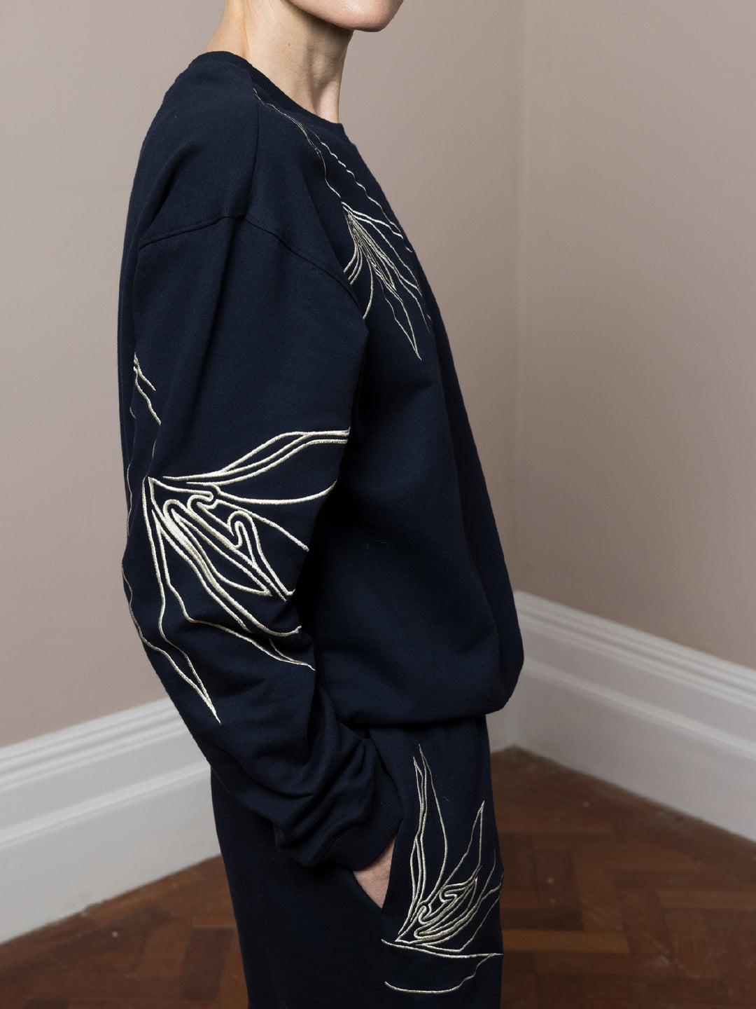 close up view view of model  standing  with hands in pockets on parquet floor wears  dark navy Joggers with artisan cream linear embroidery across pockets in cotton fleece with matching sweatshirt in dark navy with cream artisan linear embroidery across front and arms 
