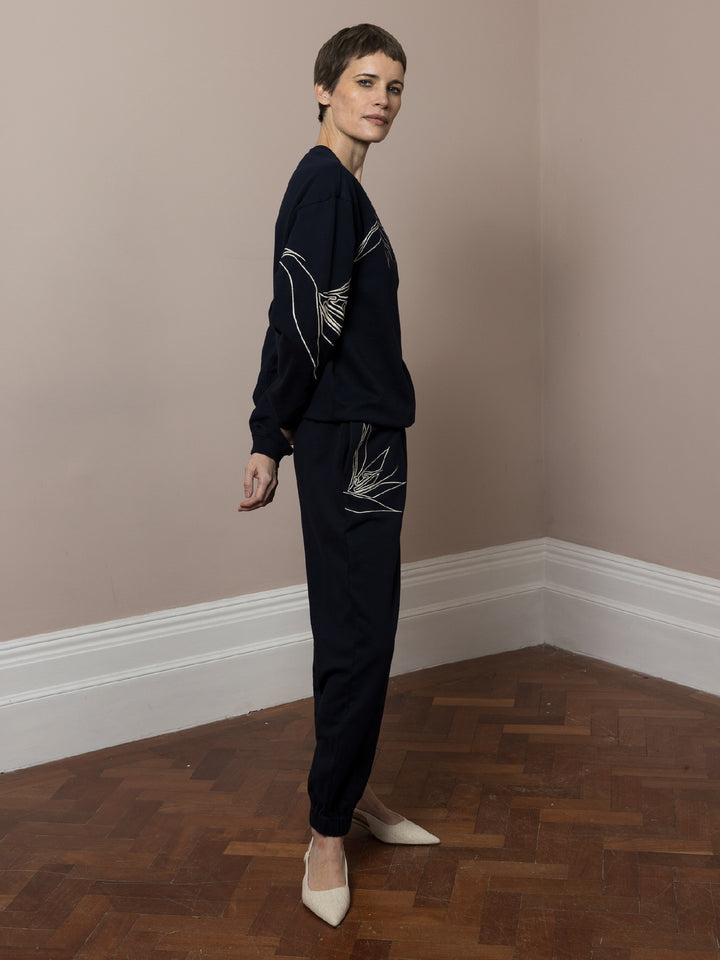 side view of model  standing  with hands in pockets on parquet floor wears  dark navy Joggers with artisan cream linear embroidery across pockets in cotton fleece with matching sweatshirt in dark navy with cream artisan linear embroidery across front and arms 